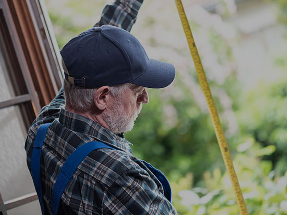 Tradesman wearing hat and plaid shirt measuring window frame with tape measure.